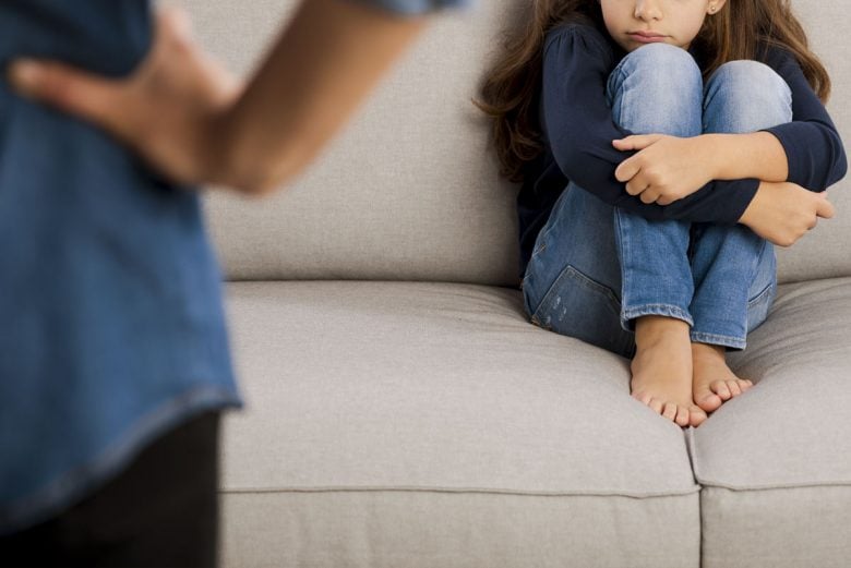 Child sitting on a couch timidly, while parent stands by them with hands on their hips.